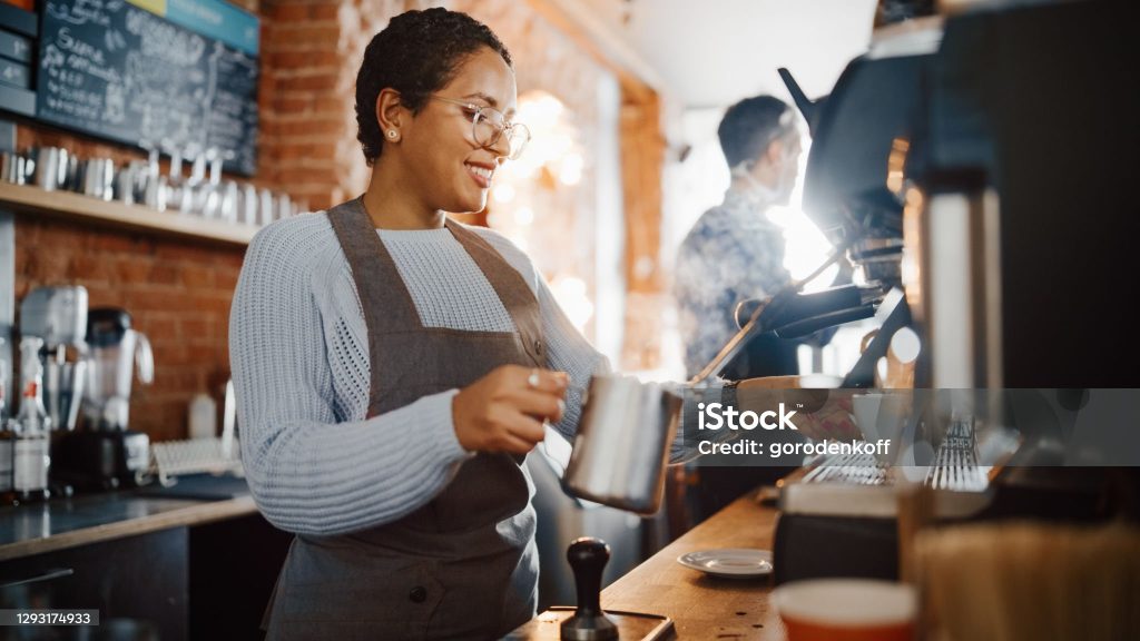 Beautiful Latin American Female Barista with Short Hair and Glasses is Making a Cup of Tasty Cappuccino in Coffee Shop Bar. Portrait of Happy Employee Behind Cozy Loft-Style Cafe Counter.